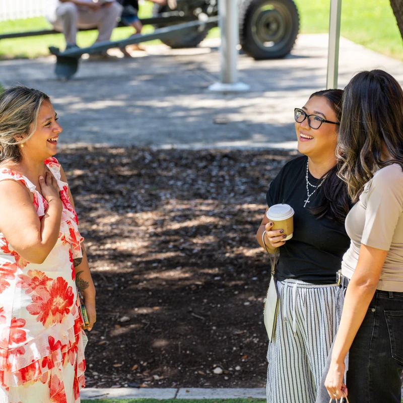 Three women stand together outside on a sunny day, laughing and talking. The woman in the center holds a coffee cup, and a large tree is beside them.
