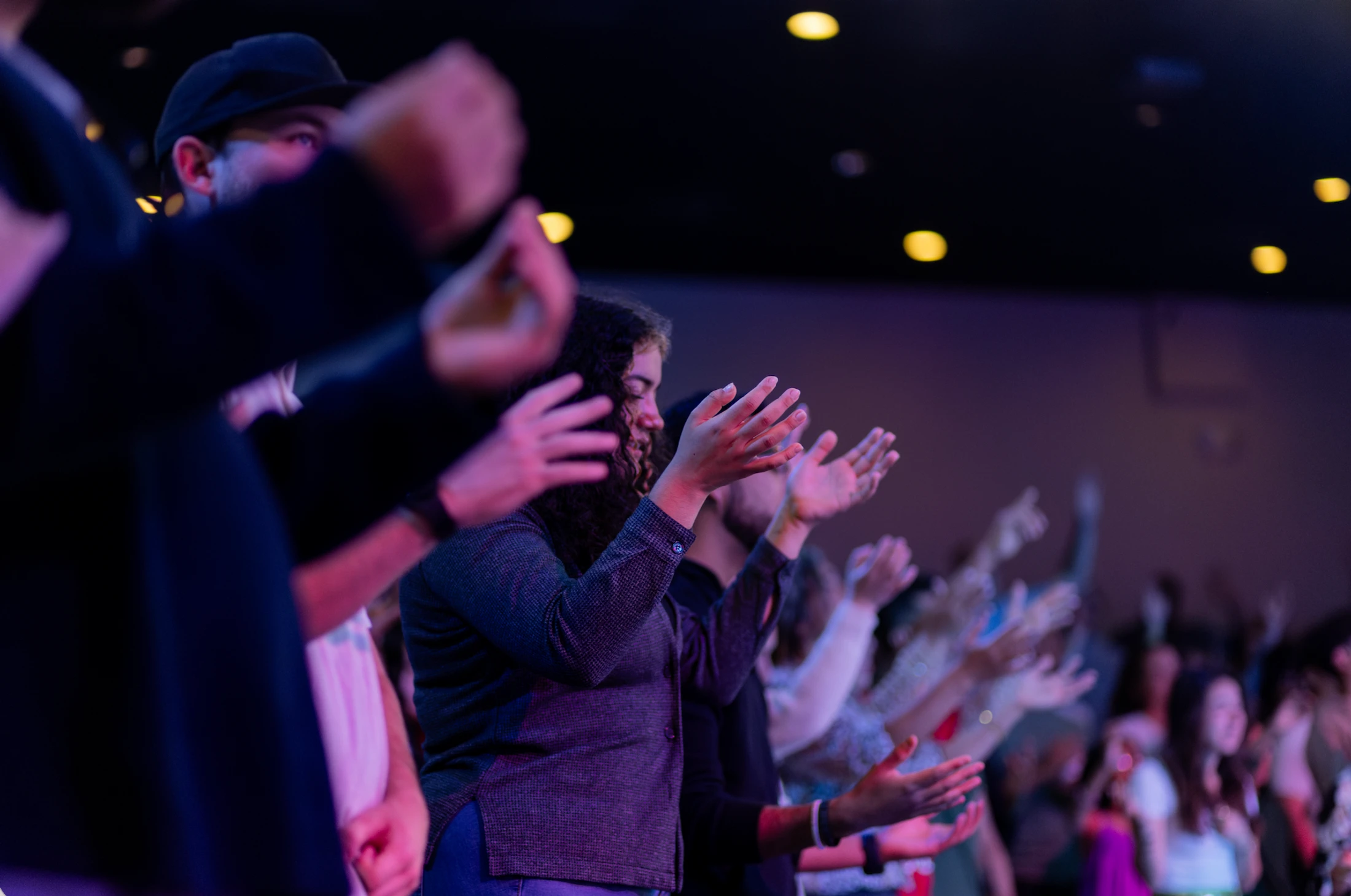 A group of people in a dimly lit auditorium with purple and blue lights, raising their hands in an act of worship. The focus is on a woman in the center.