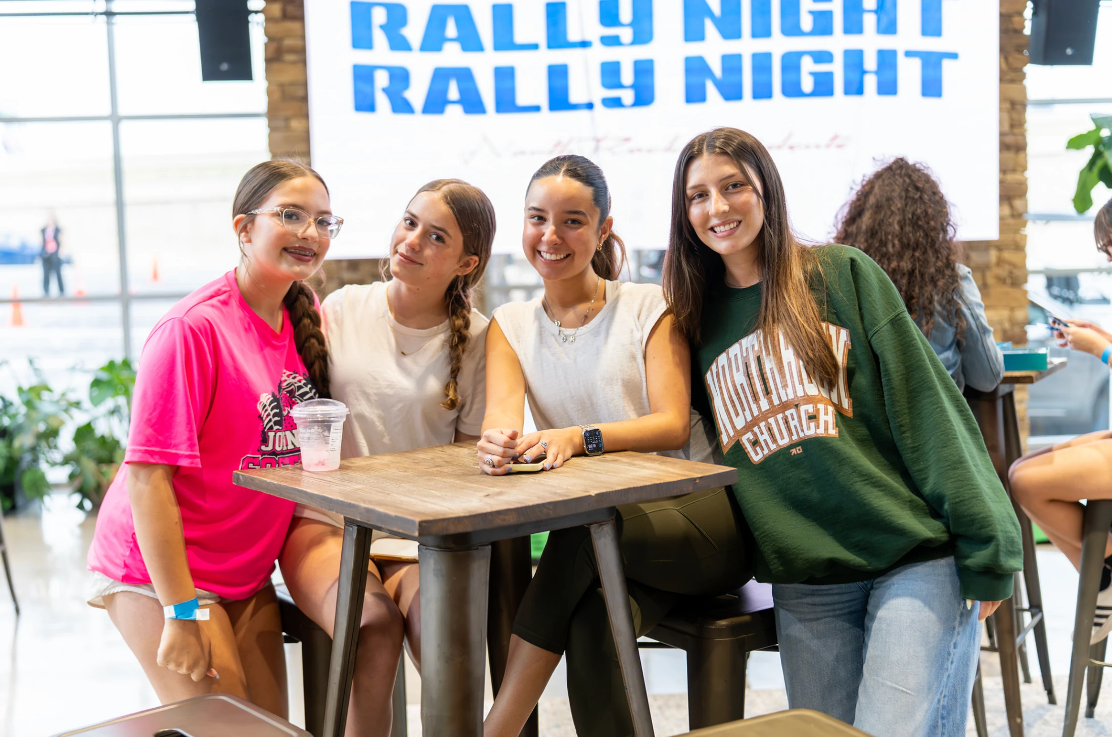 Four young women smile for a photo while sitting at a tall wooden table indoors, with a sign for "Rally Night" visible in the background.