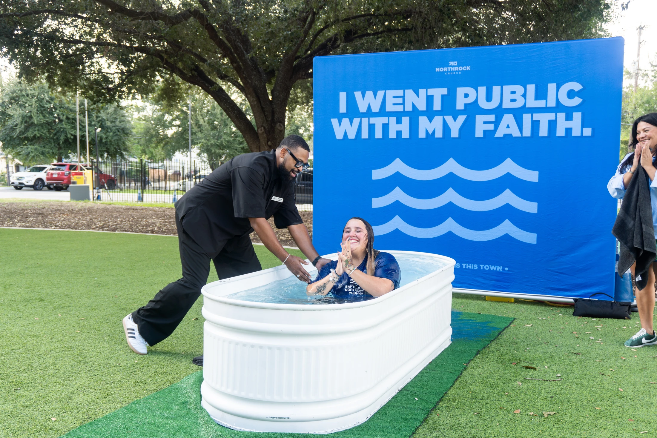 A smiling woman is baptized by a man in a white tub outdoors, with a blue banner behind them reading 'I went public with my faith.'