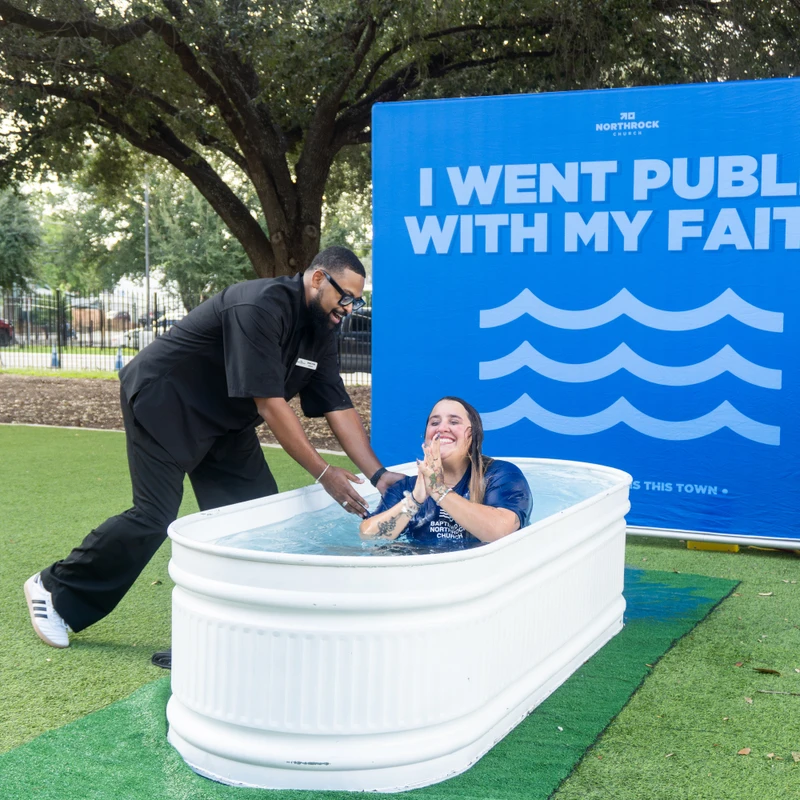 A smiling woman is baptized by a man in a white tub outdoors, with a blue banner behind them reading 'I went public with my faith.'