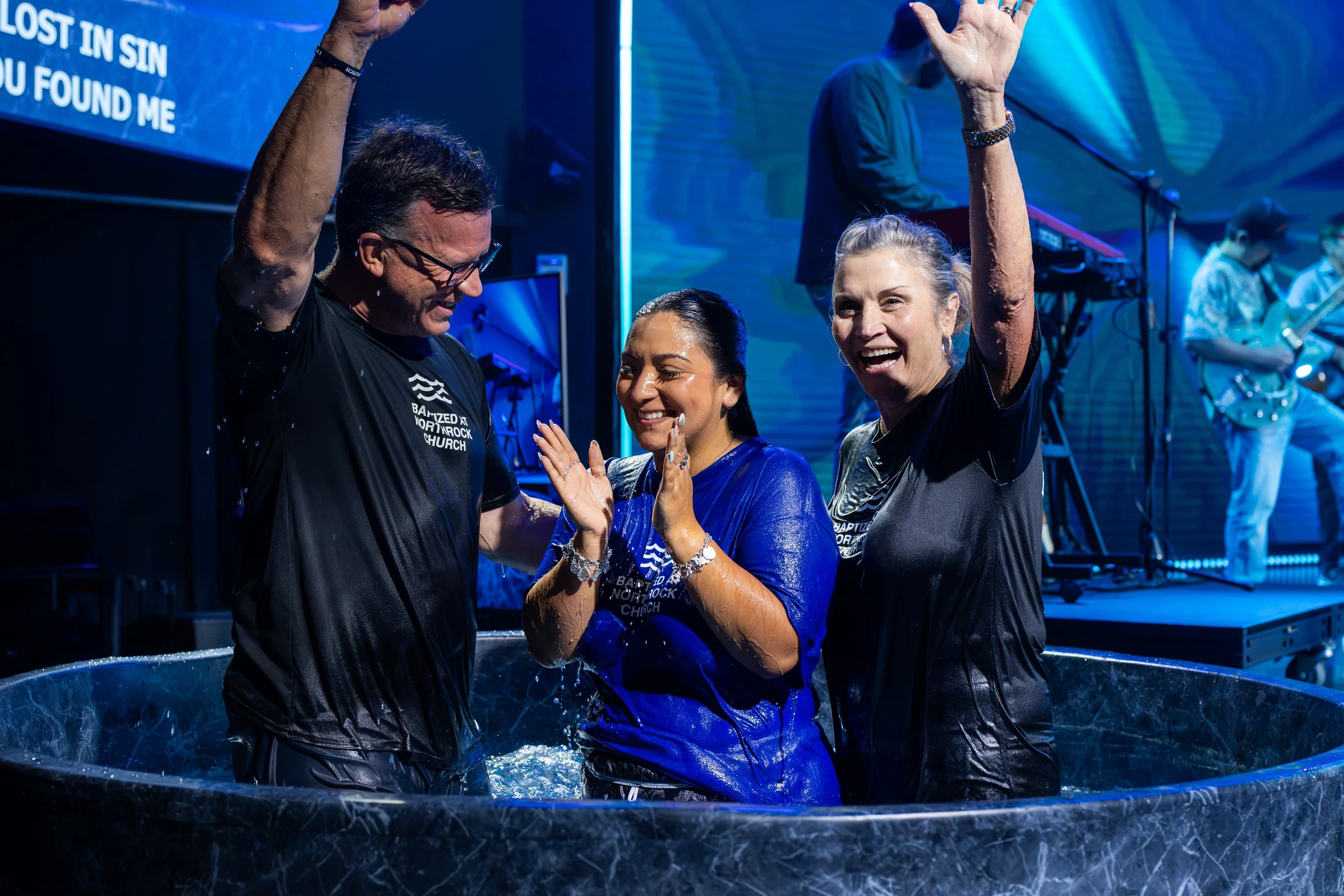 A woman in a blue shirt celebrates with a man and another woman in a baptismal font on a stage, all smiling with their hands raised in joy.
