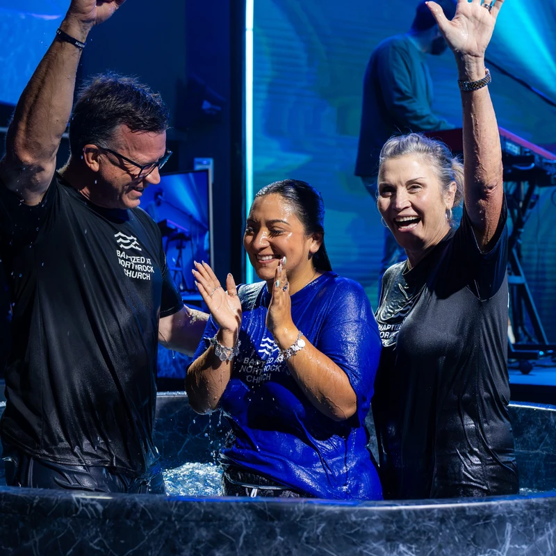 A woman in a blue shirt celebrates with a man and another woman in a baptismal font on a stage, all smiling with their hands raised in joy.