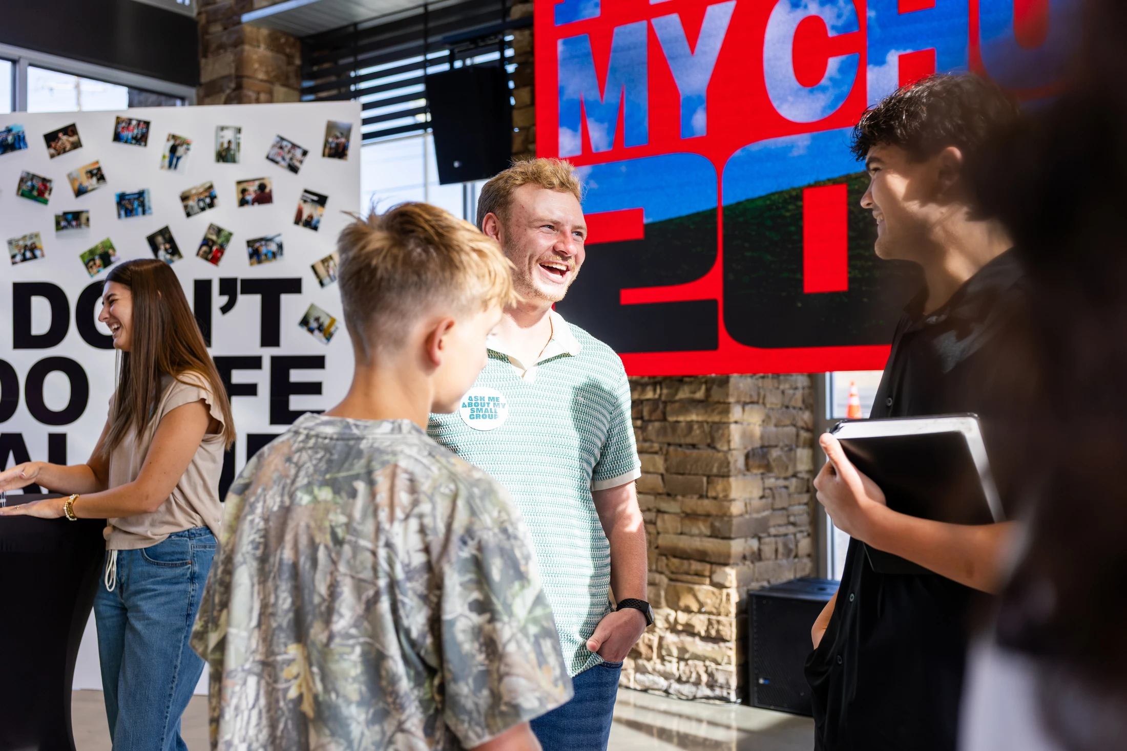 A smiling man wearing a green polo and a button that reads 'Ask Me About My Small Group' talks with two young men in a bright, modern lobby.