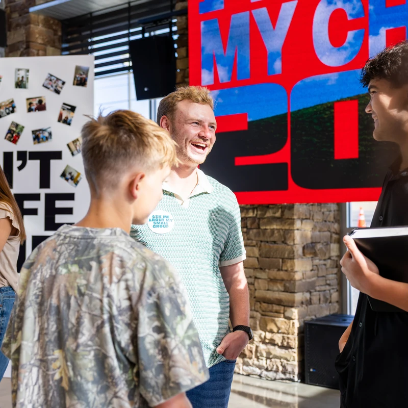 A smiling man wearing a green polo and a button that reads 'Ask Me About My Small Group' talks with two young men in a bright, modern lobby.
