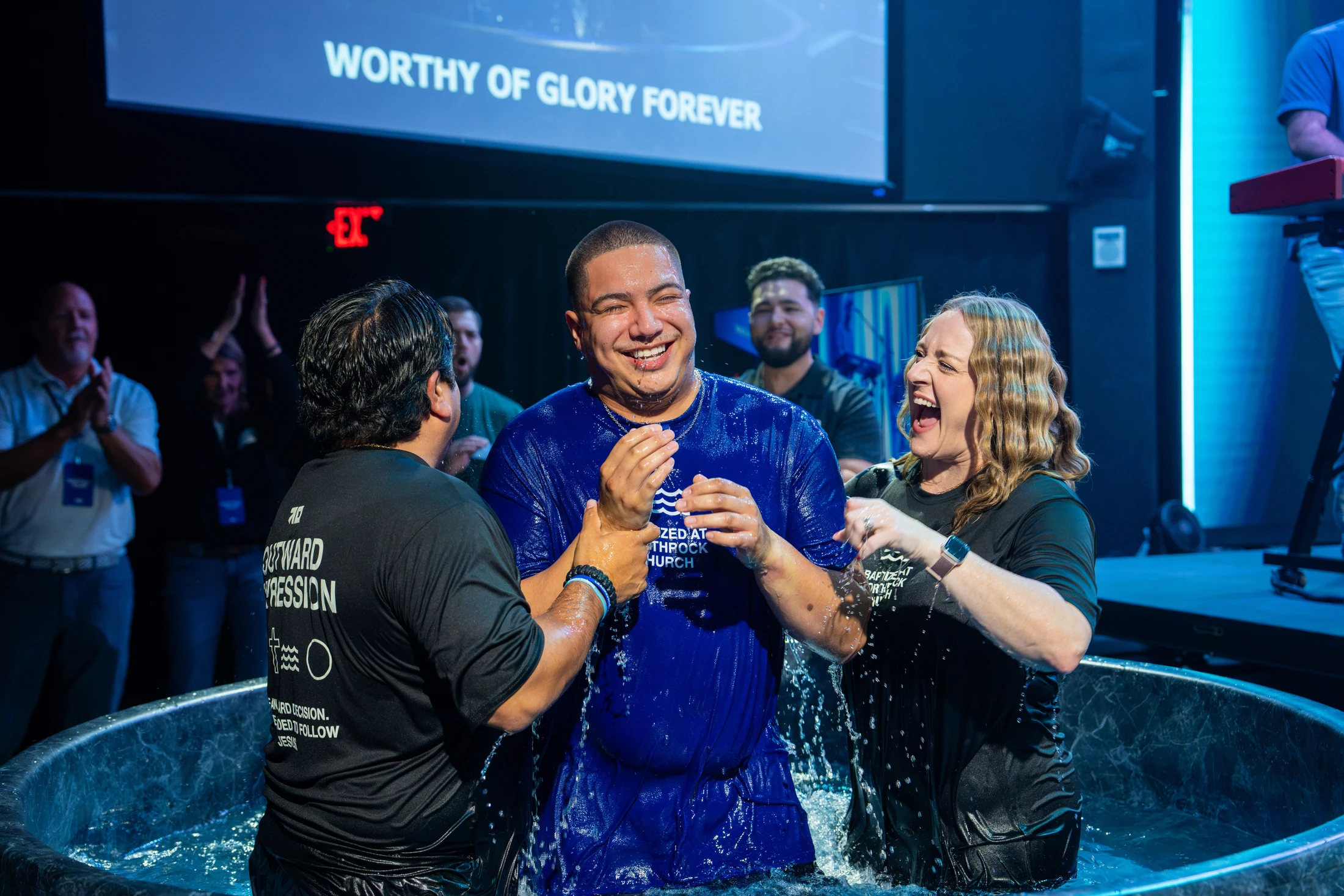 A joyful man in a wet blue shirt emerges from a baptismal pool, supported by a man and a laughing woman, during a church ceremony on a stage.