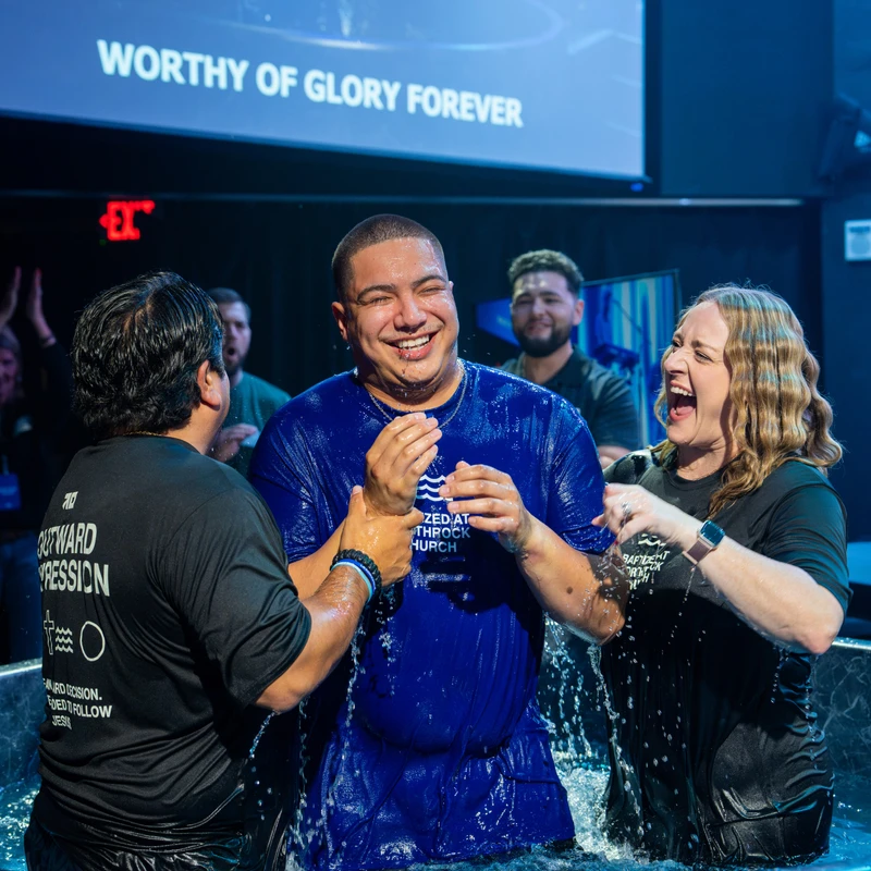 A joyful man in a wet blue shirt emerges from a baptismal pool, supported by a man and a laughing woman, during a church ceremony on a stage.