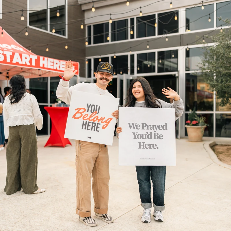A smiling man and woman stand outside a building holding welcome signs. The man's says 'You Belong Here' and the woman's says 'We Prayed You'd Be Here.'