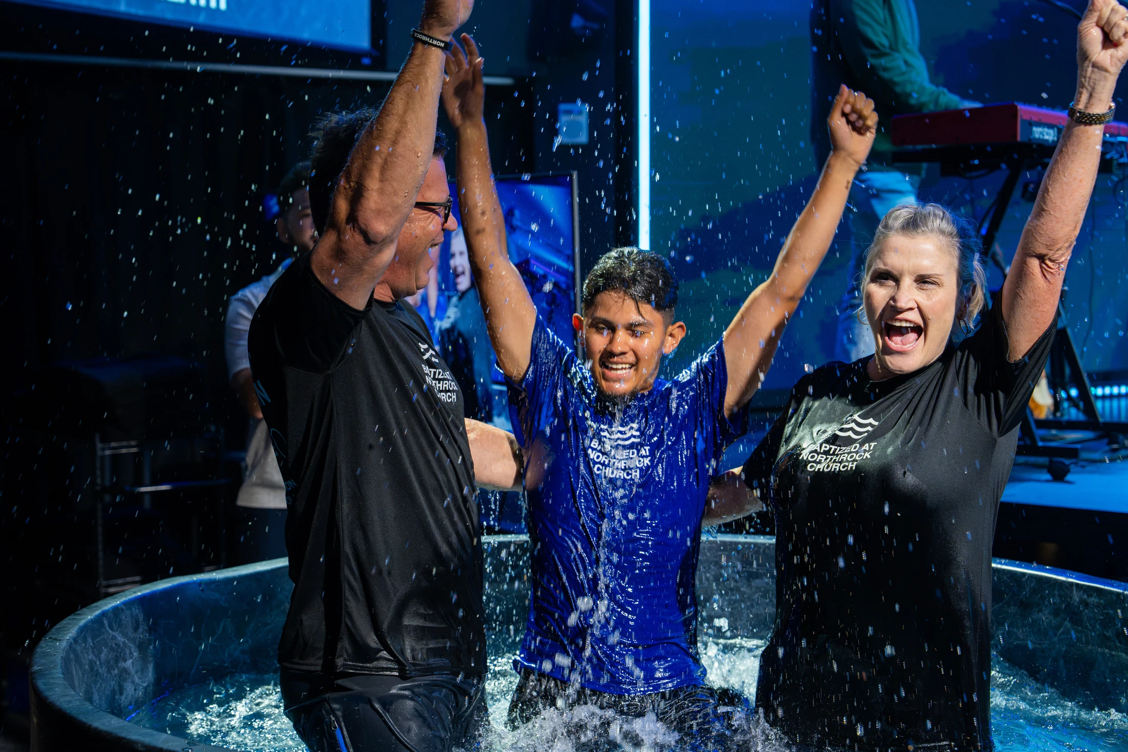 A young man raises his arms in celebration as he emerges from the water of a baptismal pool, flanked by a man and woman who are also celebrating.