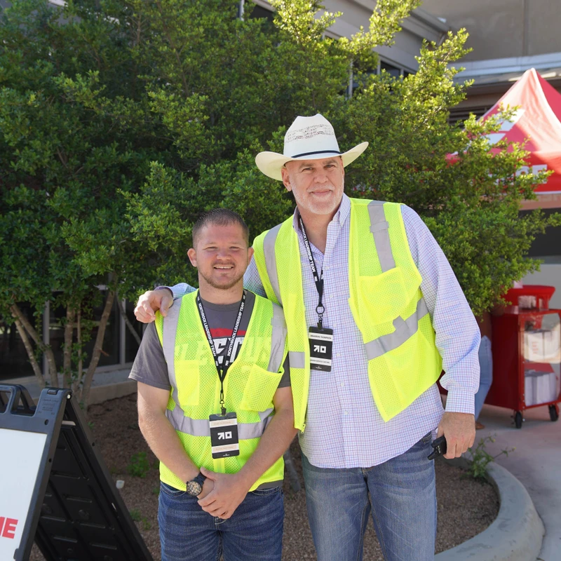 Two men in yellow safety vests and jeans smile for a photo outdoors. The taller man wears a white cowboy hat and has his arm around the other man's shoulder.