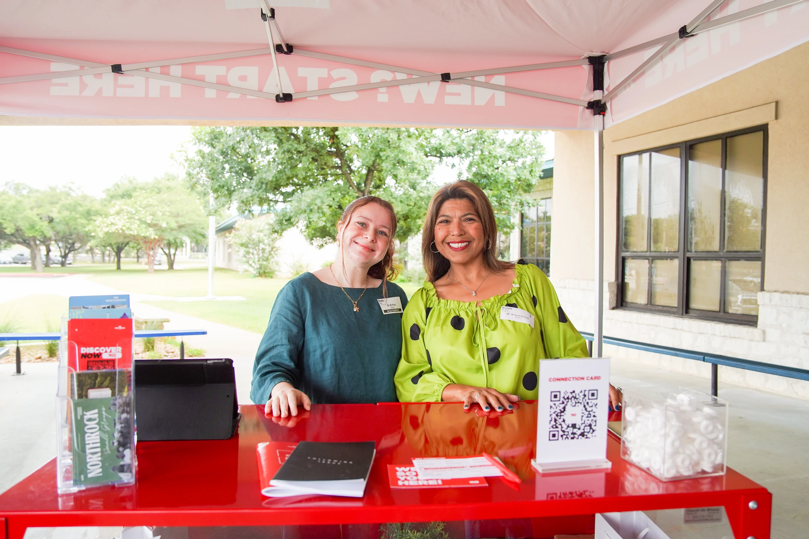 Two women smile while standing behind a red information table under a tent that reads "NEW? START HERE!" outdoors, ready to greet visitors.