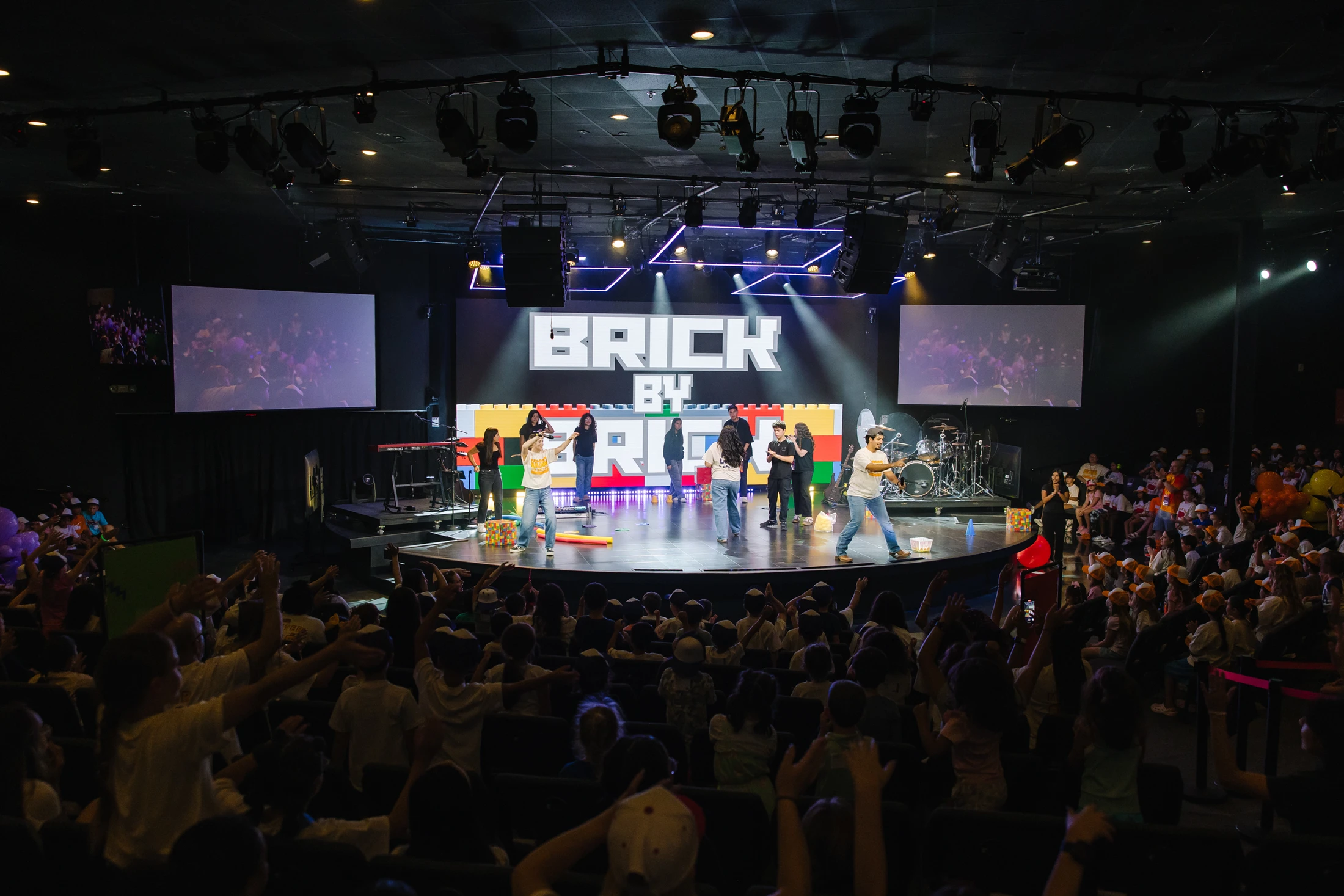 A wide shot of an auditorium filled with children watching a live performance on stage, with a large screen reading 'BRICK BY' in the background.