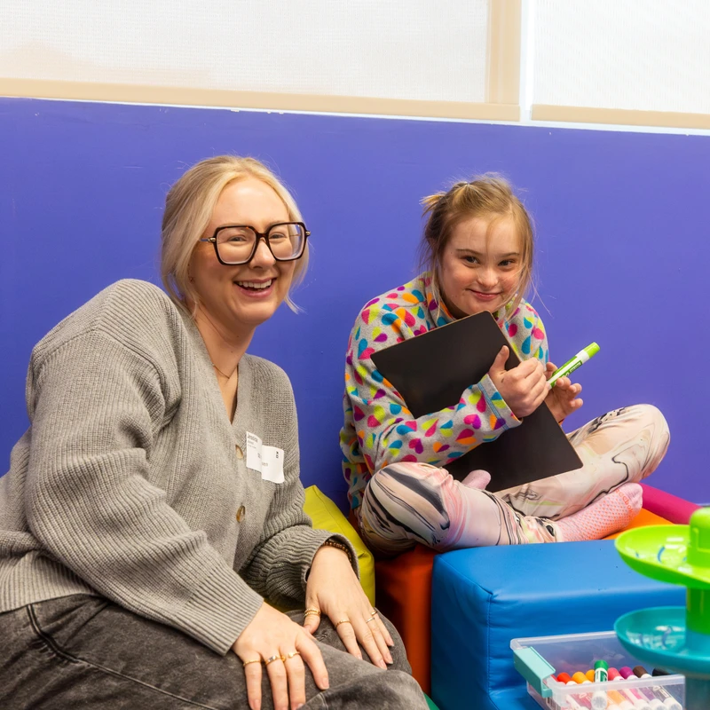 A smiling woman with glasses sits on the floor next to a happy young girl with Down syndrome who is holding a small whiteboard and a marker.