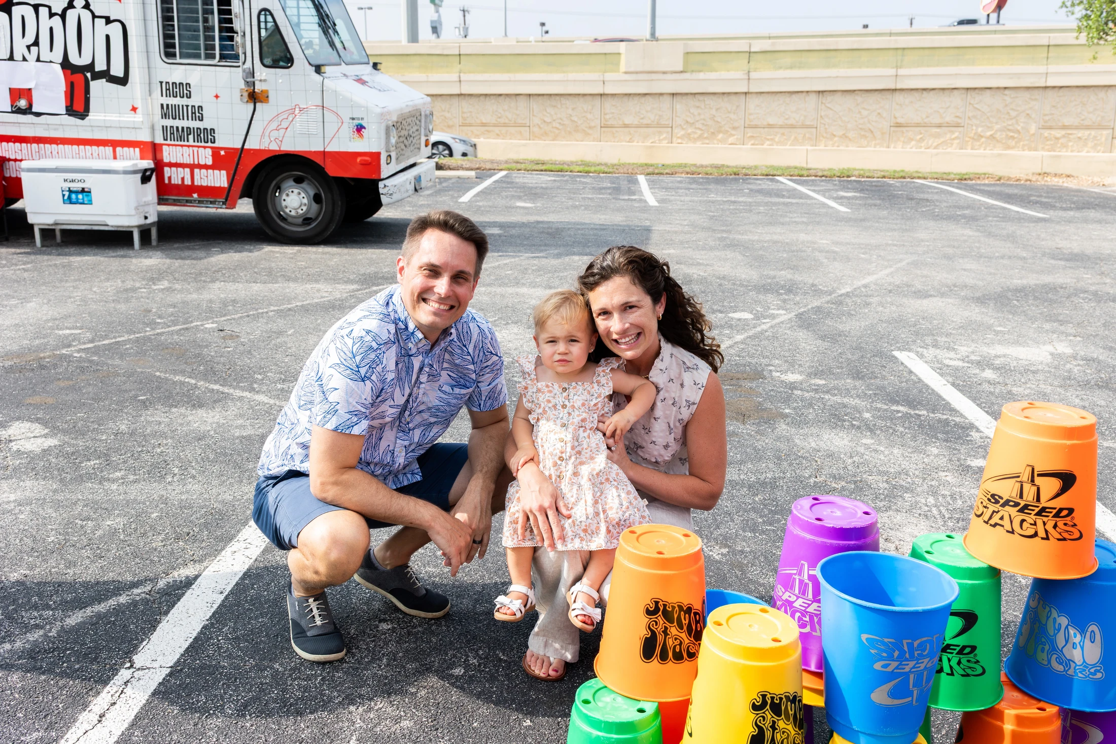 A man, woman, and toddler smile for the camera in a parking lot, with large colorful stacking cups in the foreground and a taco truck in the background.