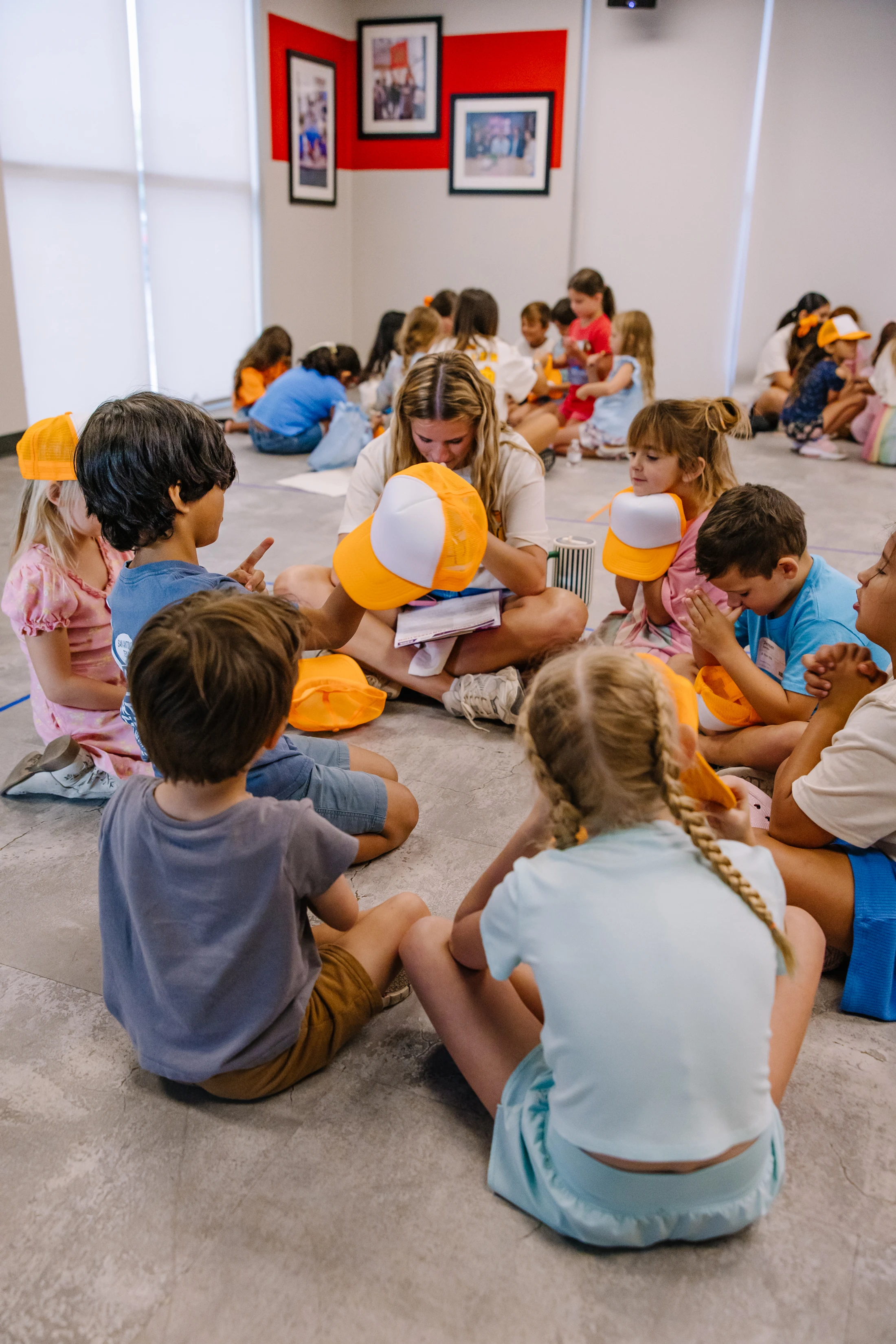 A female instructor sits cross-legged in a circle with a group of young children. They are all on the floor, holding orange and white hats during a lesson.