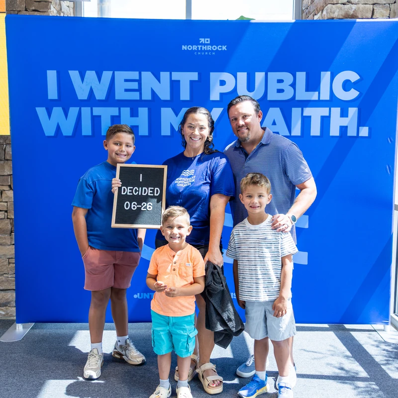 A family of five poses in front of a blue backdrop that says "I Went Public With My Faith." A boy holds a sign that reads "I Decided 06-26."