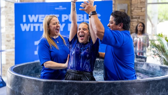 A woman joyfully raises her arms in celebration as she emerges from a baptismal pool, flanked by a smiling man and woman who are assisting her.
