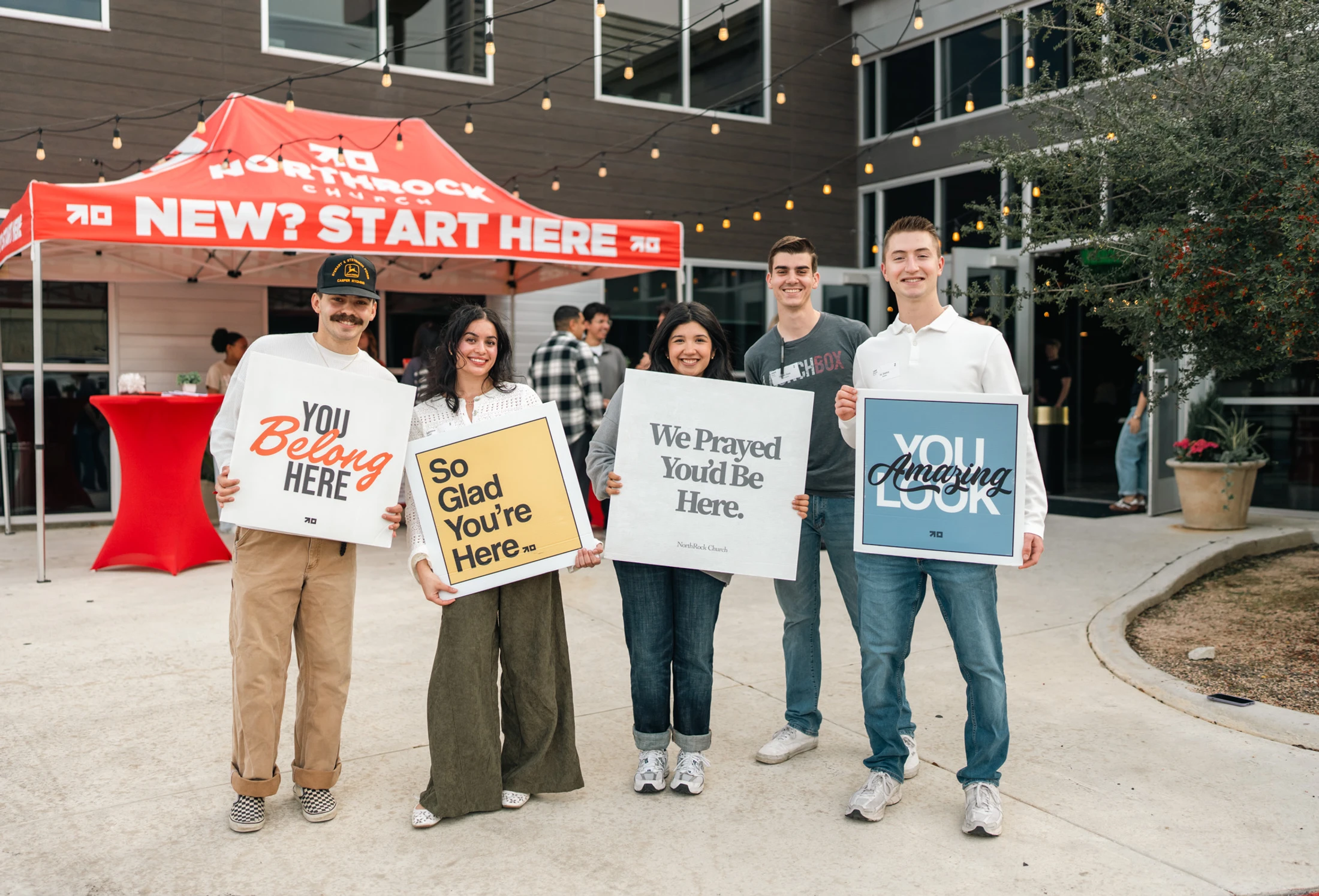 Five young adults from a church welcome team stand outside holding signs with messages like 'You Belong Here' and 'We Prayed You'd Be Here'.