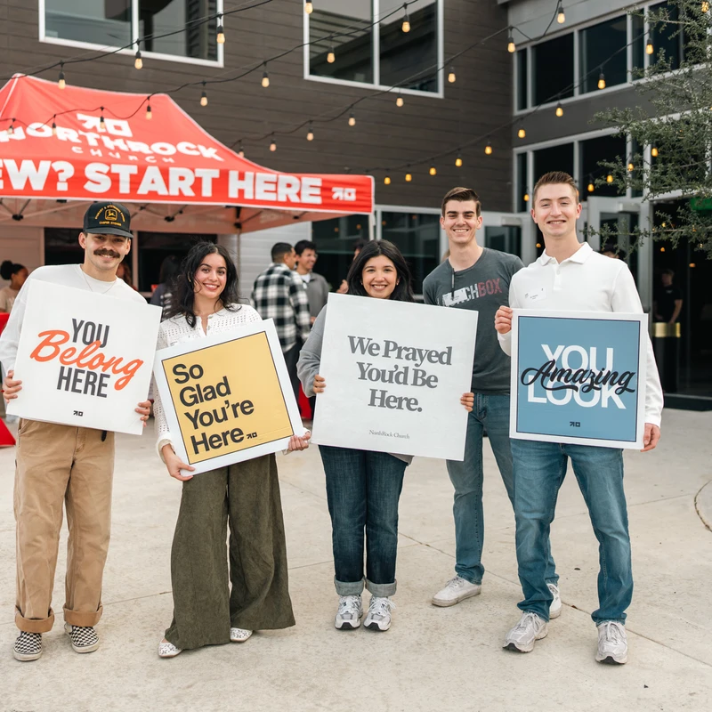 Five young adults from a church welcome team stand outside holding signs with messages like 'You Belong Here' and 'We Prayed You'd Be Here'.