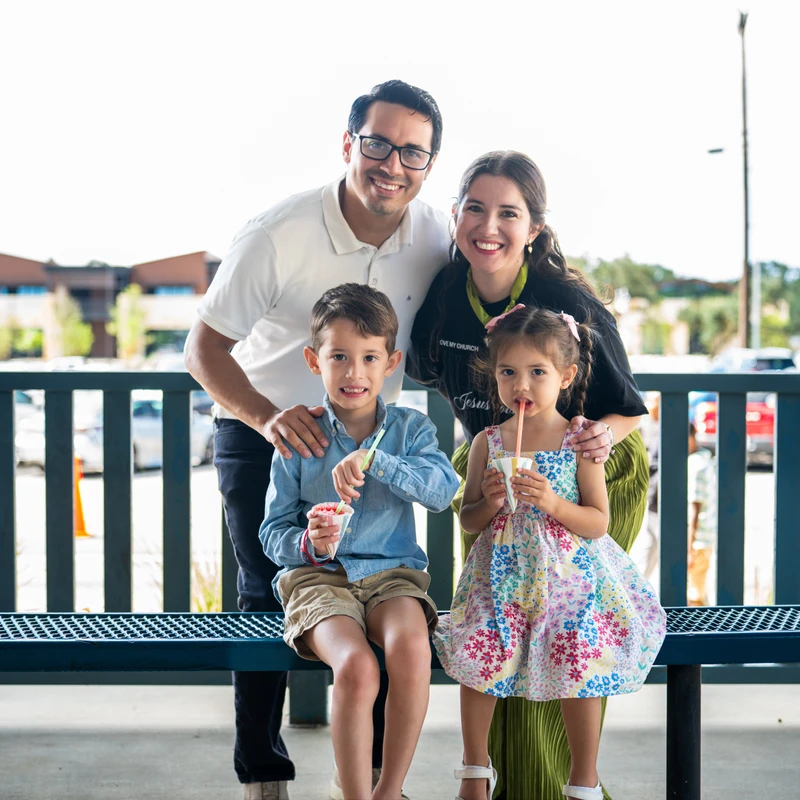 A family of four poses for a photo. The parents stand smiling behind their son and daughter, who are sitting on a blue bench eating snow cones.