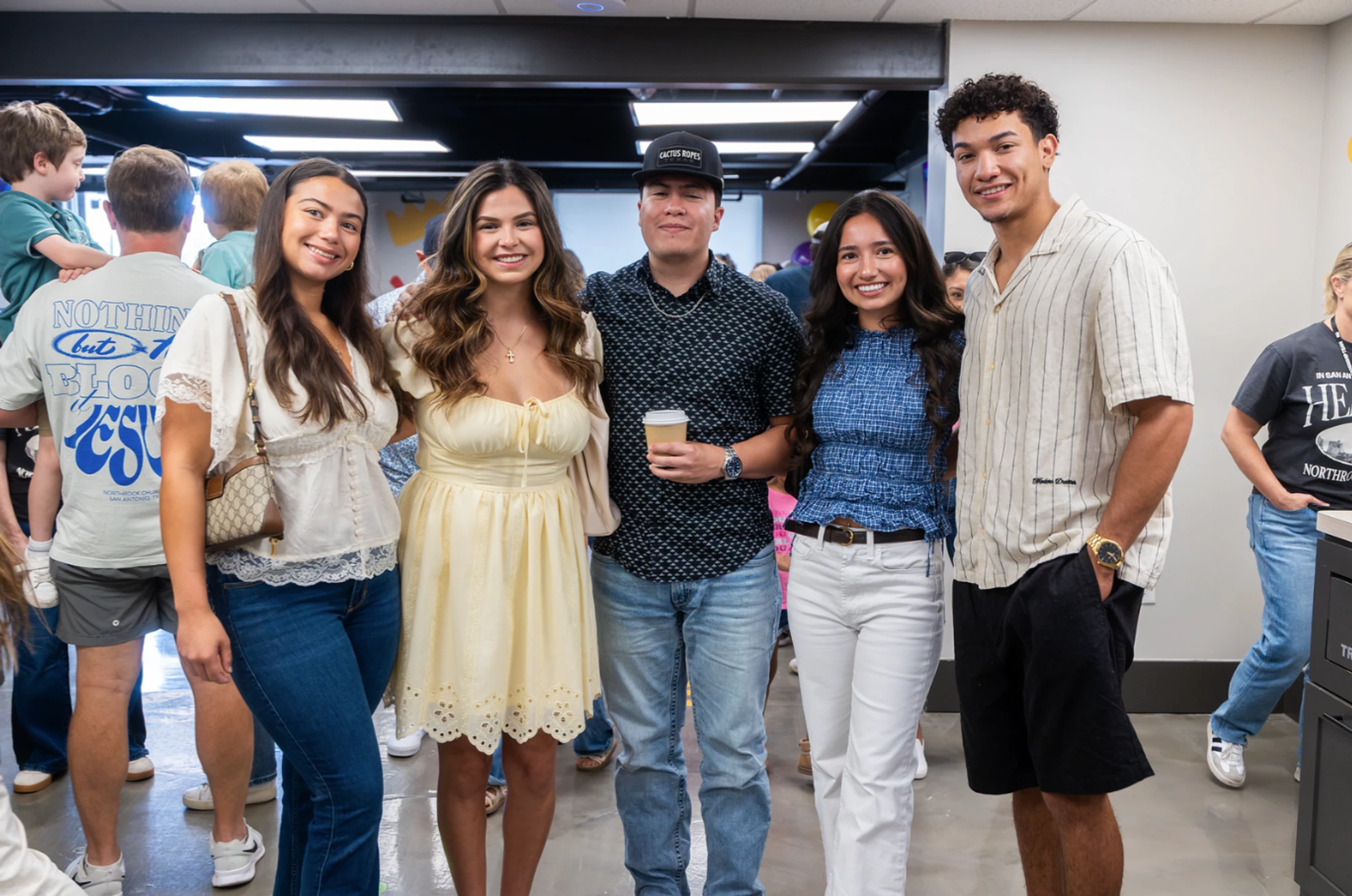 A group of five young adults, three women and two men, stand together and smile for the camera at an indoor gathering.