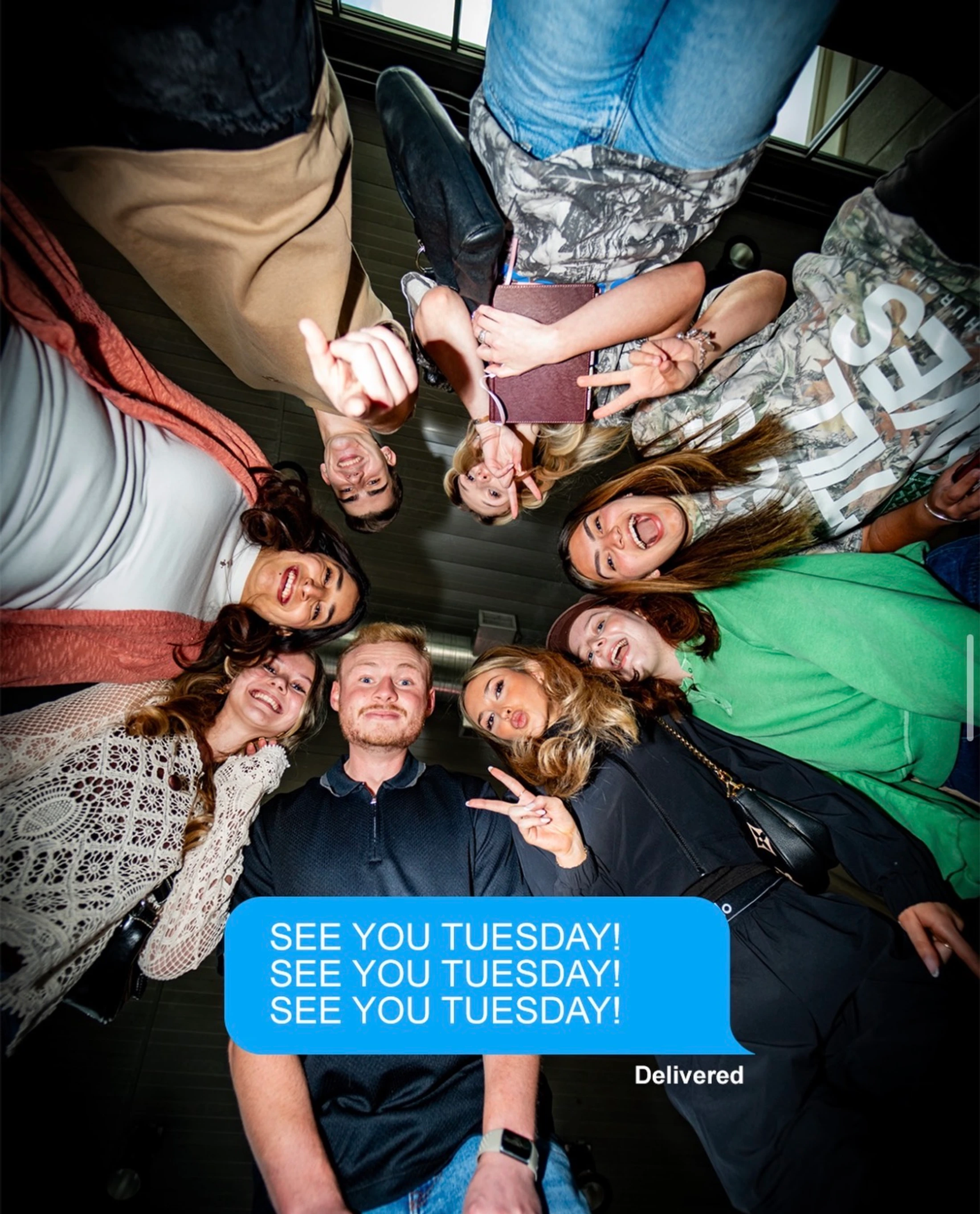 A low-angle shot of a group of young people in a circle looking down at the camera and smiling, with a 'See You Tuesday!' text message overlay.