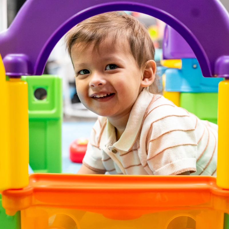 A smiling young boy with brown hair lies on his stomach, peeking happily through the purple arch of a brightly colored plastic play structure.
