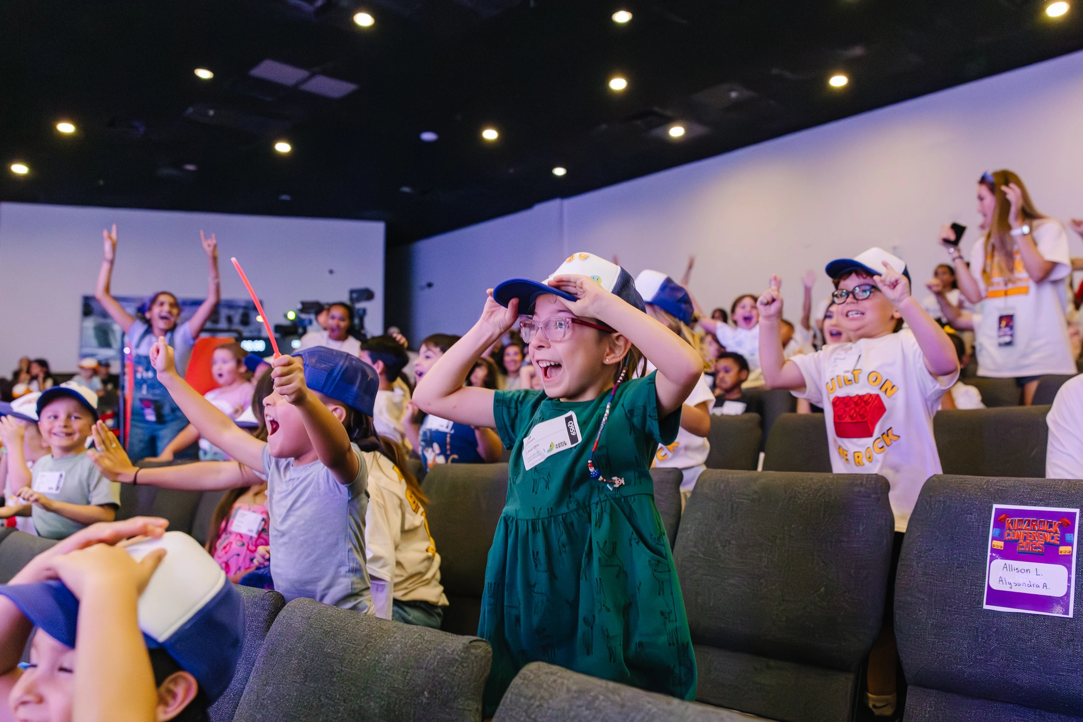 A diverse group of excited children in an auditorium stand and cheer with their hands in the air, their faces full of joy and amazement during an event.