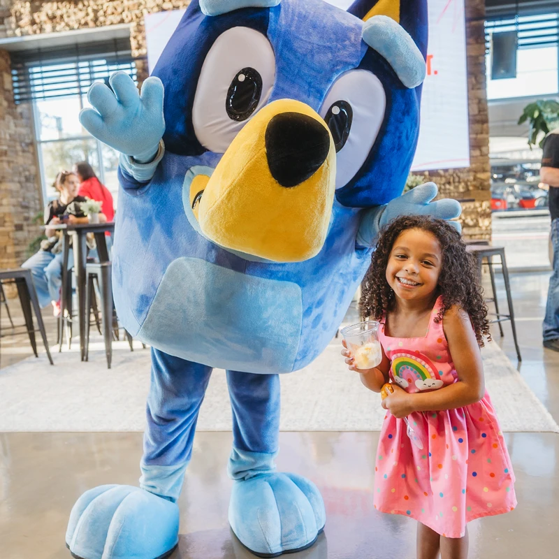 A smiling young girl in a pink rainbow dress stands next to a person in a large, blue costume of the cartoon character Bluey, who is waving.