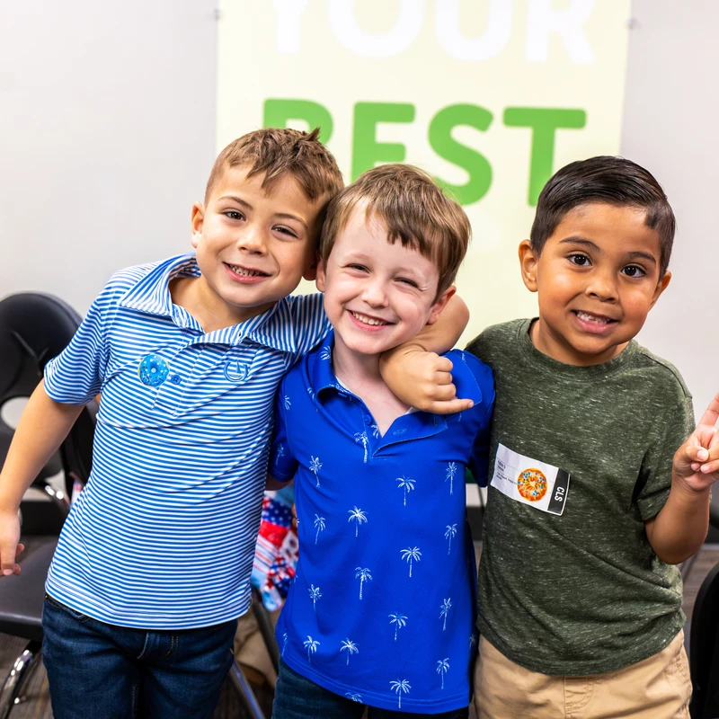 Three diverse young boys stand with their arms around each other, smiling for a photo in a classroom. The boy on the right holds up a peace sign.