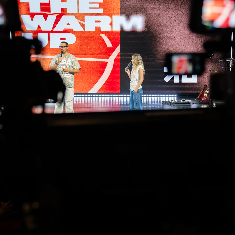 A man and a woman hold microphones on a stage in front of a large screen that reads 'THE WARM UP'. The shot is framed by out-of-focus cameras.