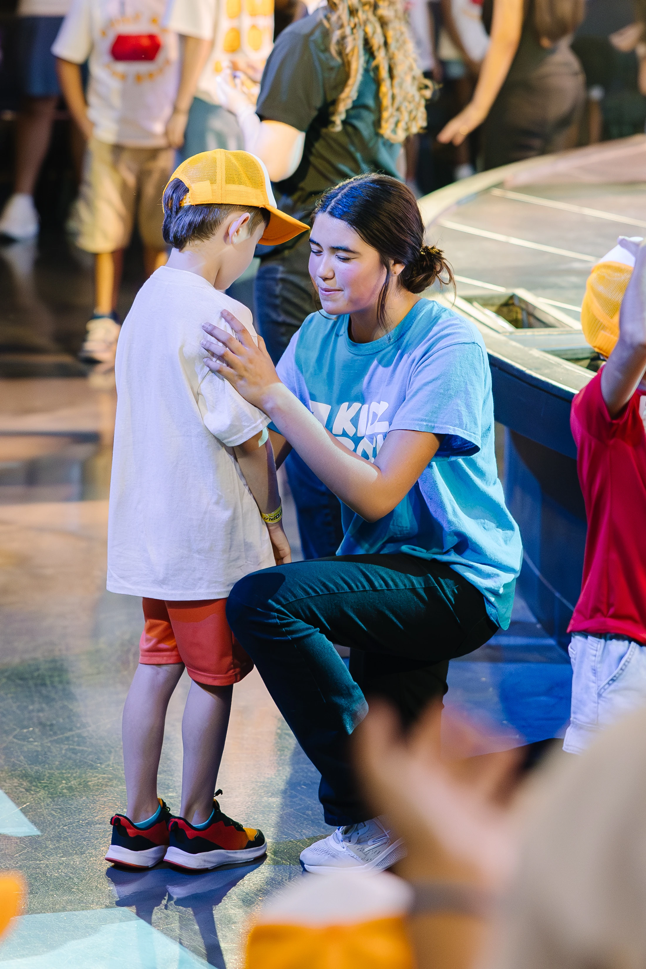 A female volunteer in a blue shirt kneels down to talk to a young boy in a yellow hat, placing her hands gently on his shoulders in a comforting gesture.