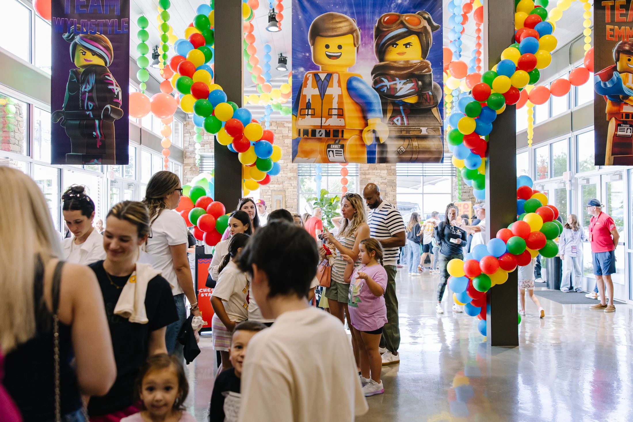 A crowded indoor hall decorated for a Lego Movie event with colorful balloon arches and large banners of the characters Emmet and Wyldstyle.