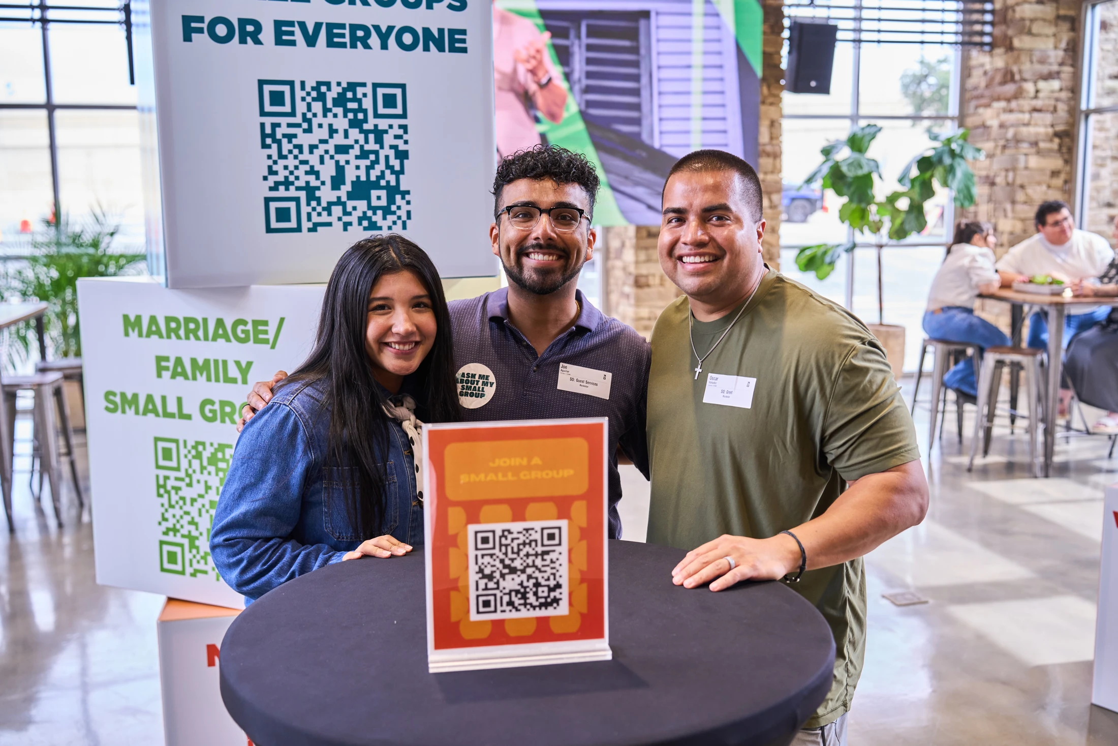 Three smiling volunteers stand behind a table with a sign and QR code to join a small group, ready to welcome people at a community event.
