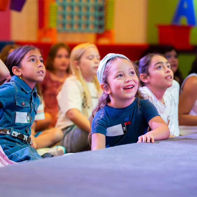 A group of young children sit on the floor, looking up with captivated expressions. A girl in a blue shirt in the foreground smiles with delight.