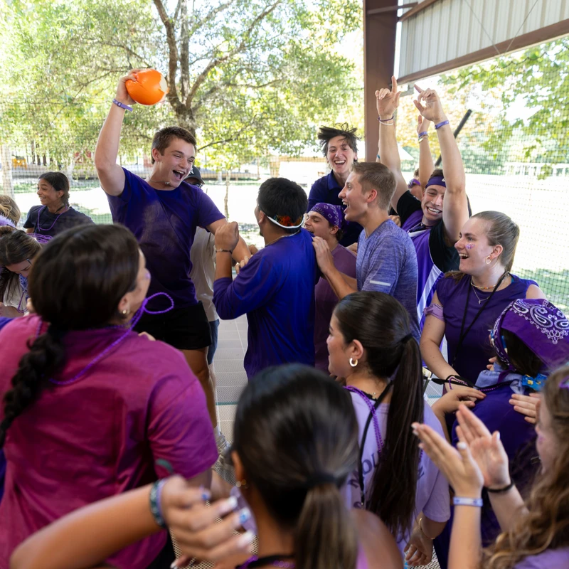A group of energetic young people in purple shirts cheering and laughing together outdoors. One boy in the center triumphantly holds an orange ball overhead.