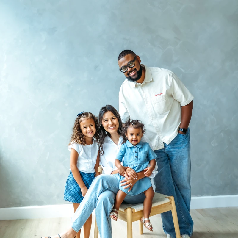 A family of four smile for a photo against a gray wall. The mother sits holding a toddler, an older daughter stands beside them, and the father stands behind.