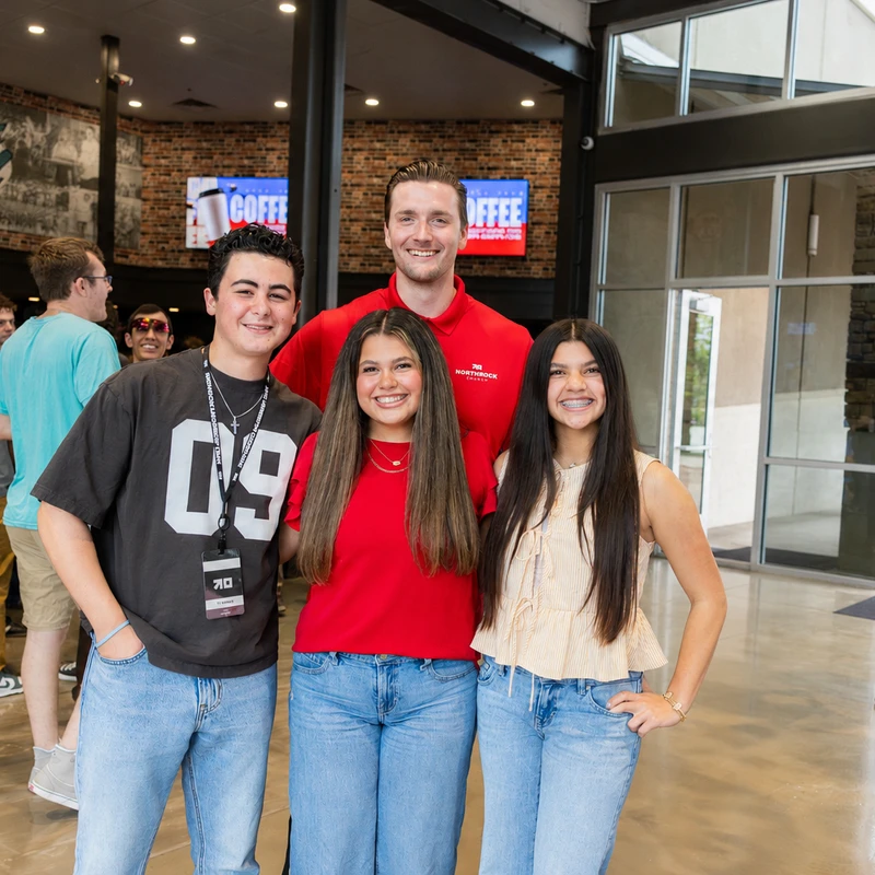 A group of three teenagers and one man smile for a photo inside a modern building's lobby, with other people mingling in the background.