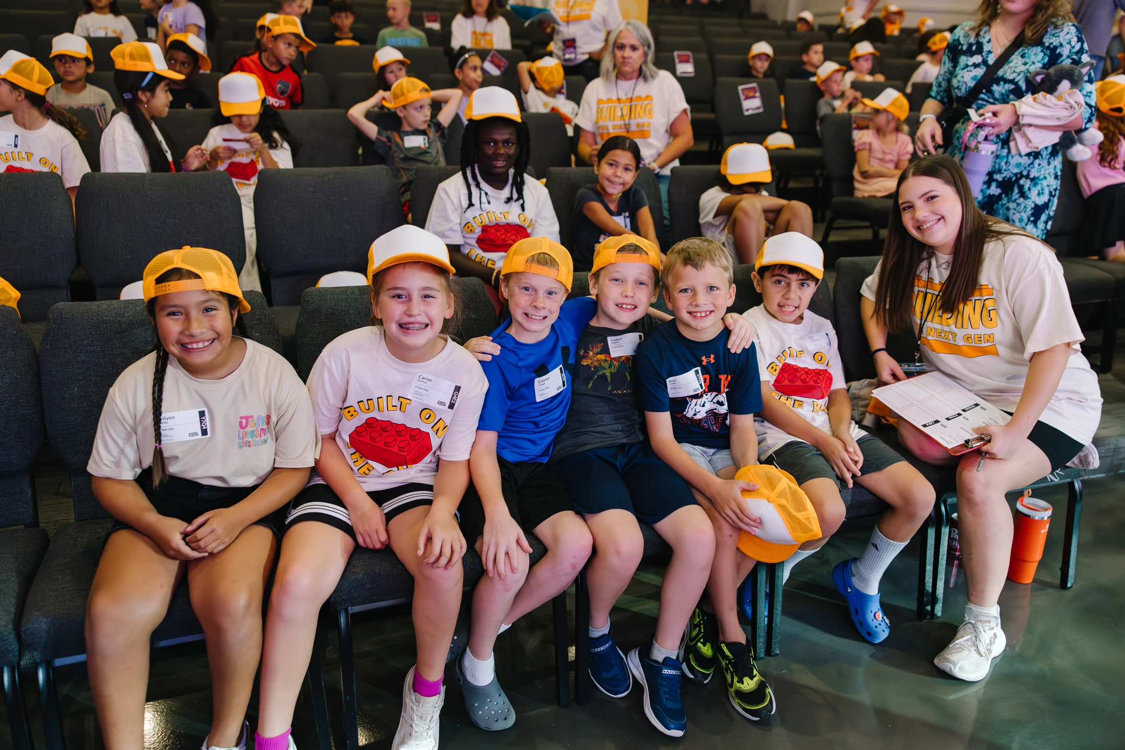 A group of six children and a young woman pose for a photo, smiling while sitting in chairs in a large room full of other kids at an event or camp.