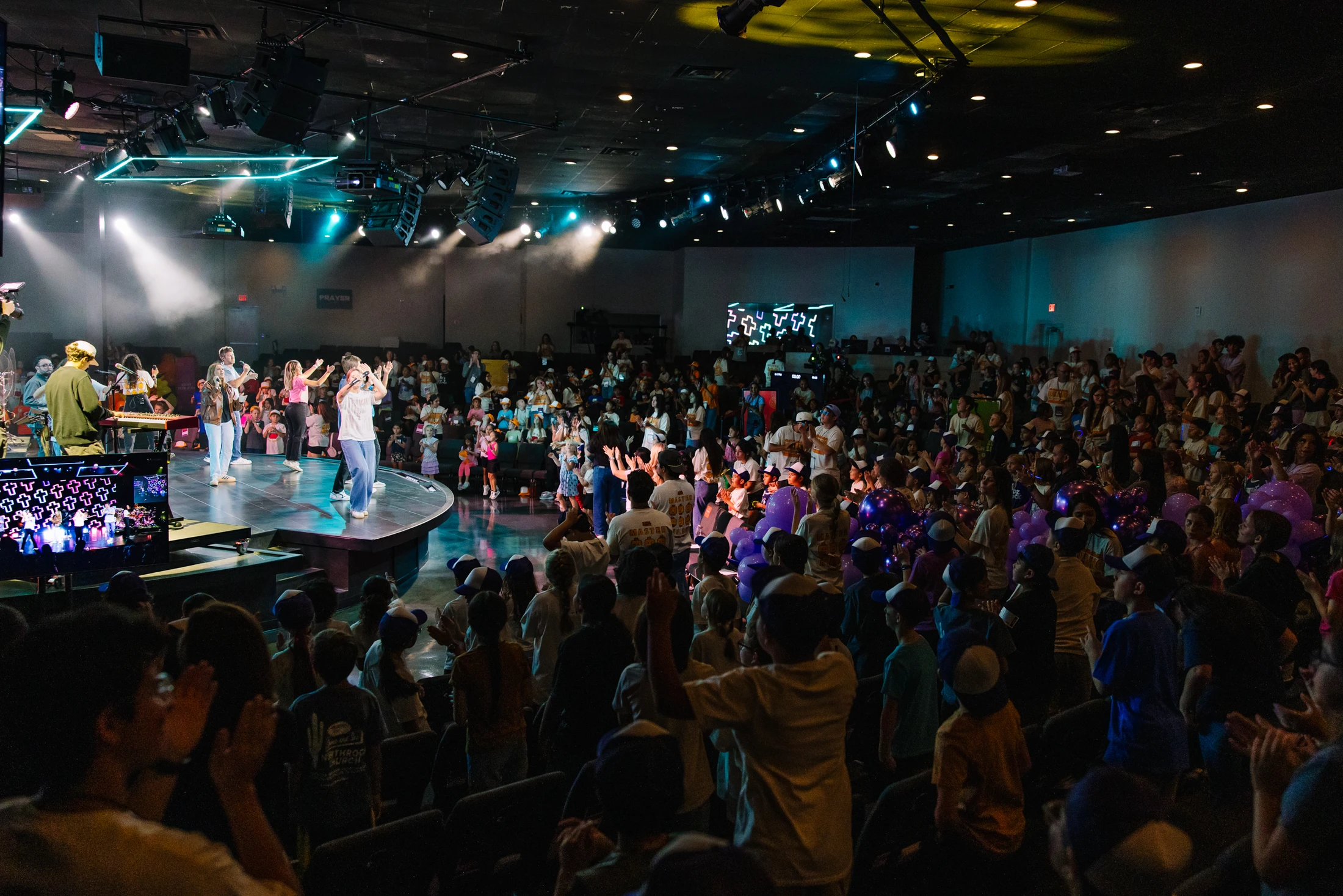 A wide shot of a crowded auditorium with a band on stage performing for an audience of children and teens, who are clapping and raising their hands.