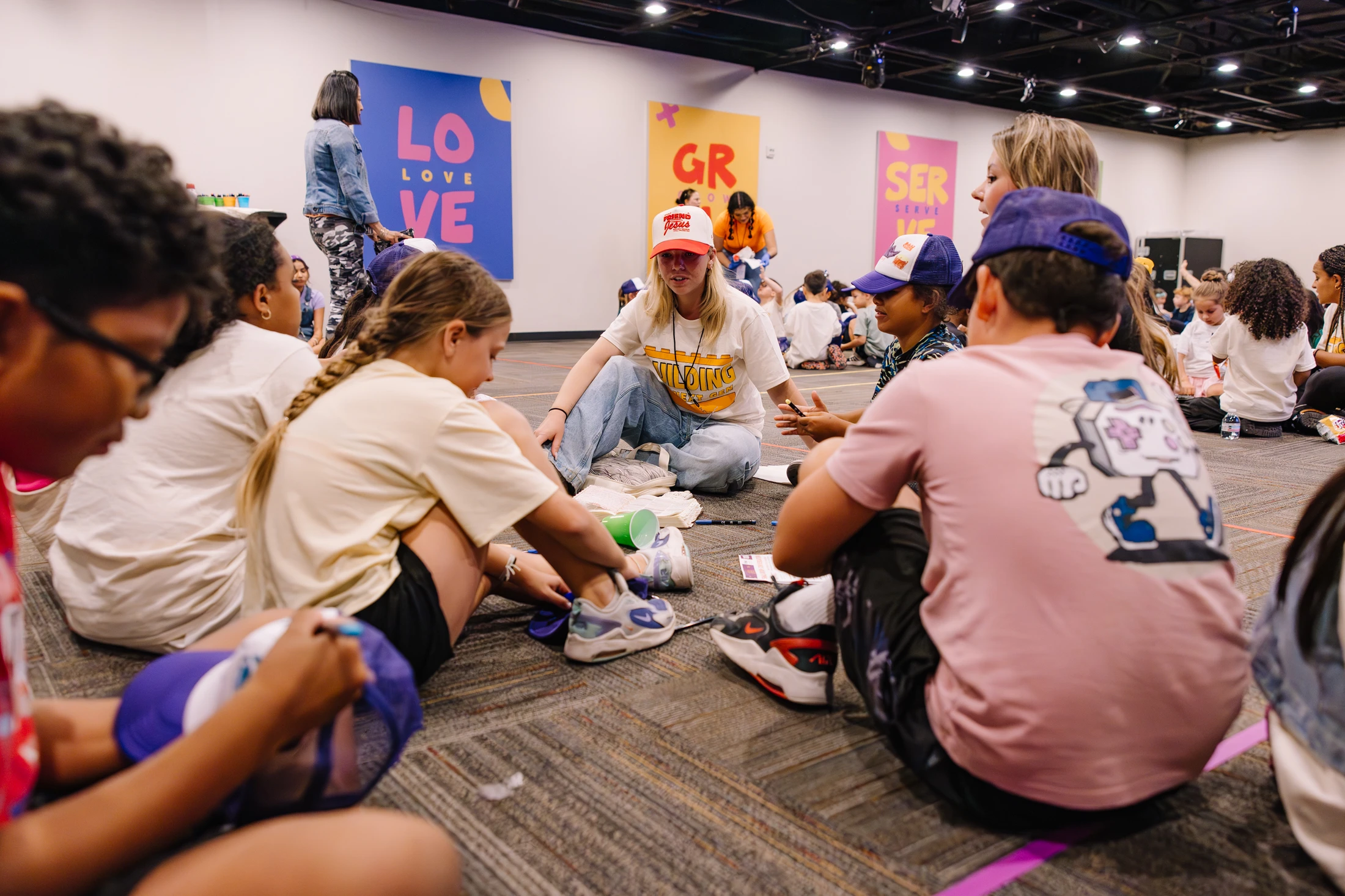 An adult leader sits on the floor in a circle with a group of diverse children, engaging them in a conversation or activity in a large event hall.