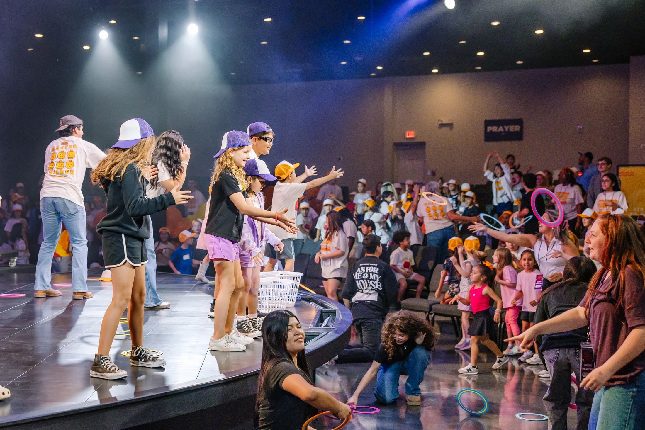 Children and adults on a brightly lit stage play a ring toss game with an enthusiastic audience during a large event in an auditorium.