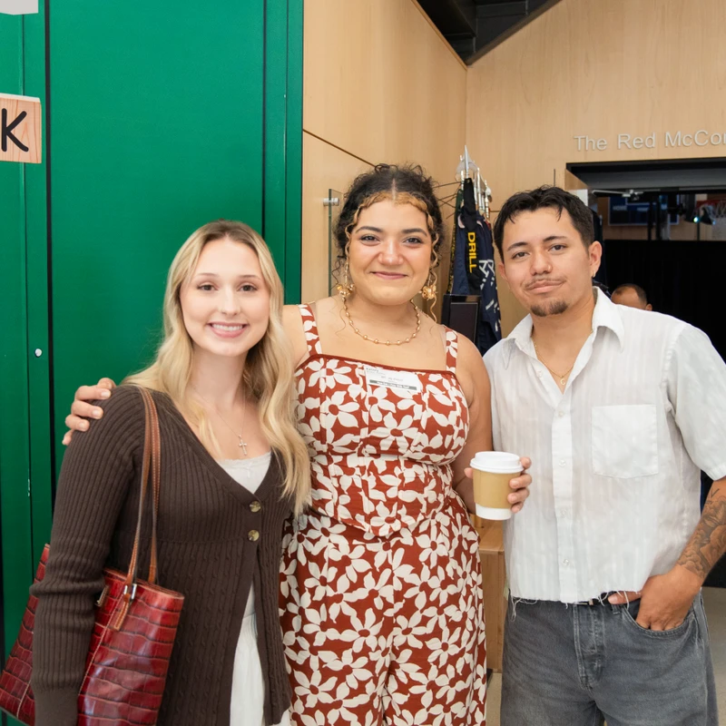 Two women and one man stand together and smile for a photo at an indoor event. In the background, a sign reads "The Red McCombs Family Gymnasium."