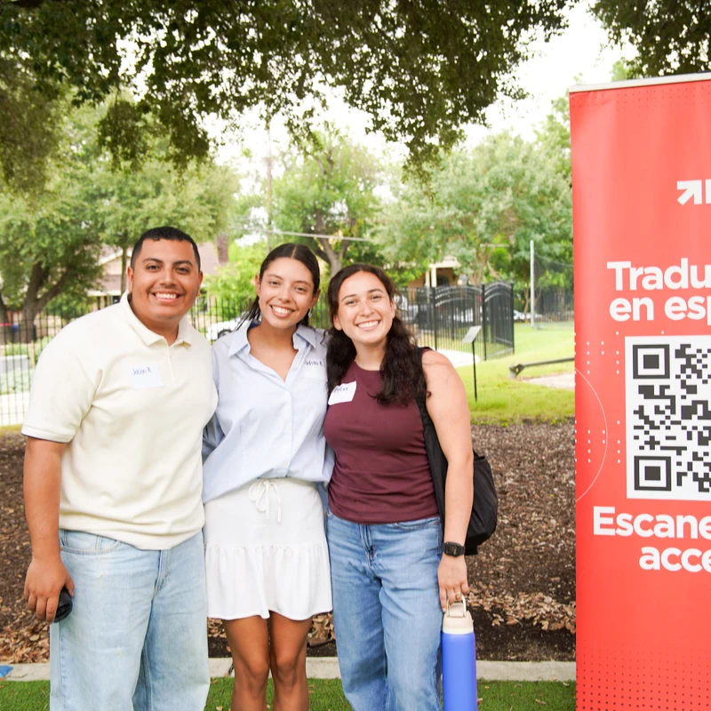 A man and two women smile for a photo outside, standing next to a red banner with a QR code and Spanish text that says 'Traducción en español'.