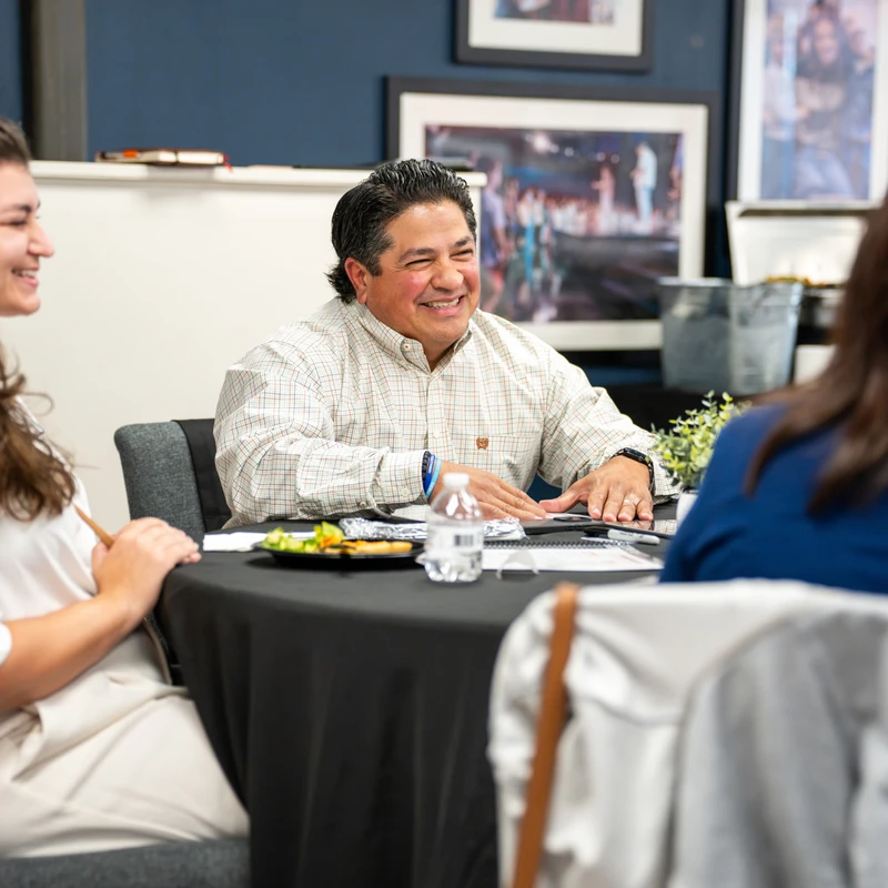 A man in a plaid shirt smiles while sitting at a round table with two women during a meeting. They appear to be engaged in a pleasant conversation.