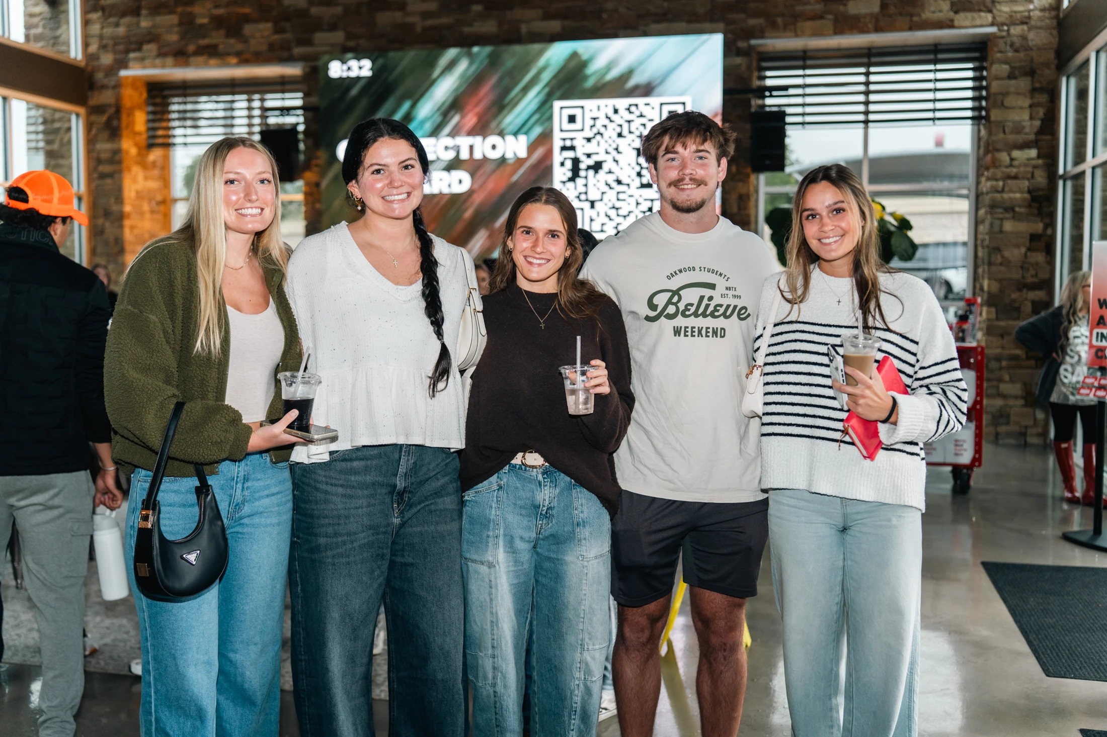 A group of five young adults, four women and one man, stand together smiling in a modern building lobby. They are dressed casually and some are holding drinks.