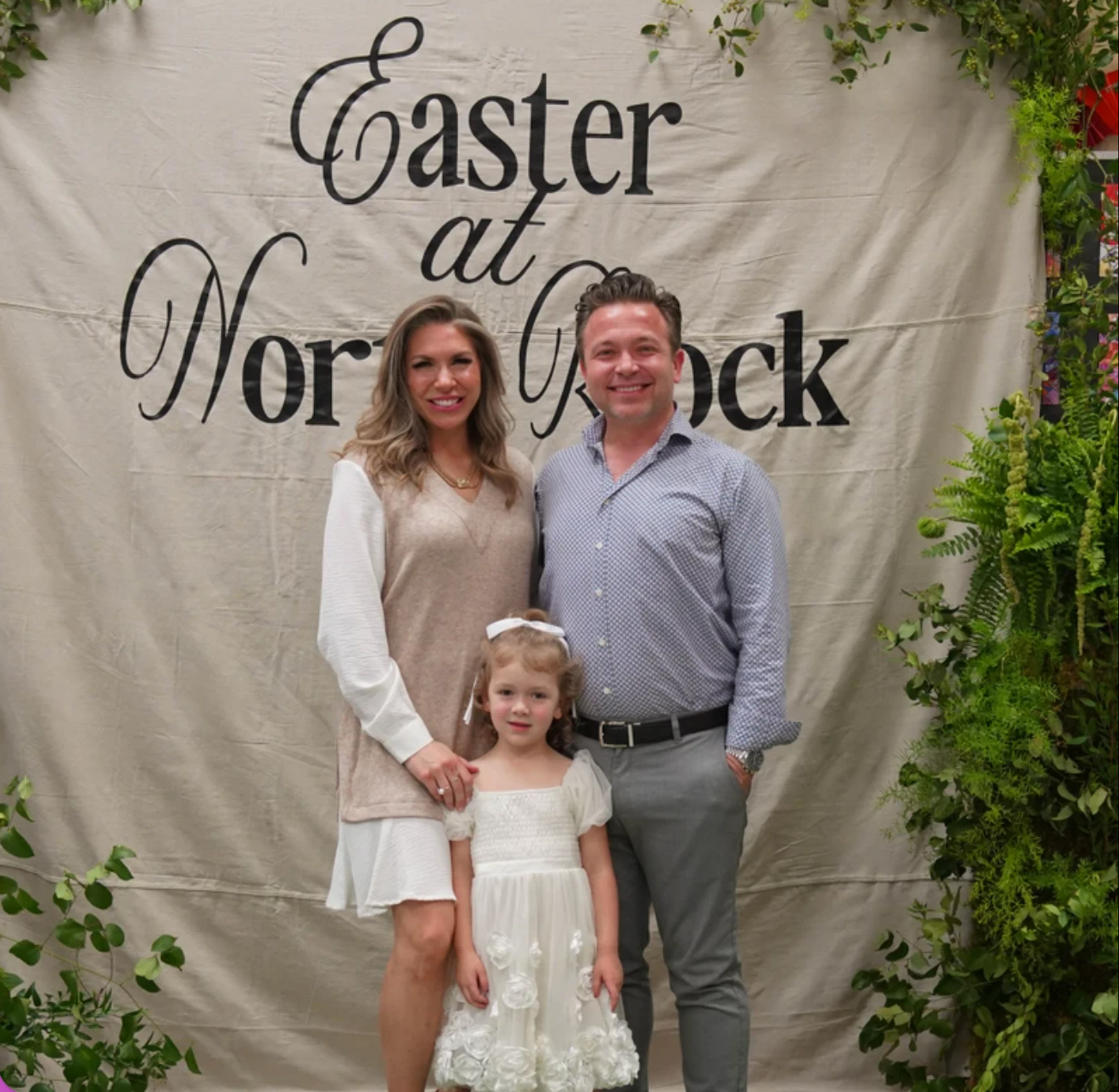 A smiling family of three, a man, woman, and young girl, pose in front of a backdrop that reads "Easter at North Rock" and is framed with greenery.