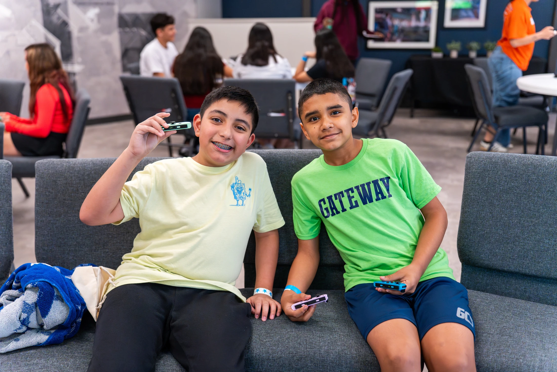 Two young boys sit on a gray couch, smiling at the camera while holding video game controllers. The boy on the left wears a yellow shirt; the other wears green.