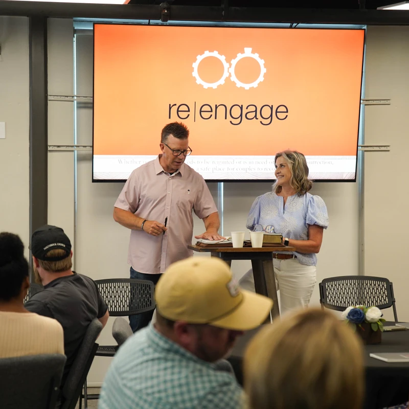 A man and a woman stand behind a table leading a workshop. Behind them, a large orange screen displays the 're|engage' logo with two gears.
