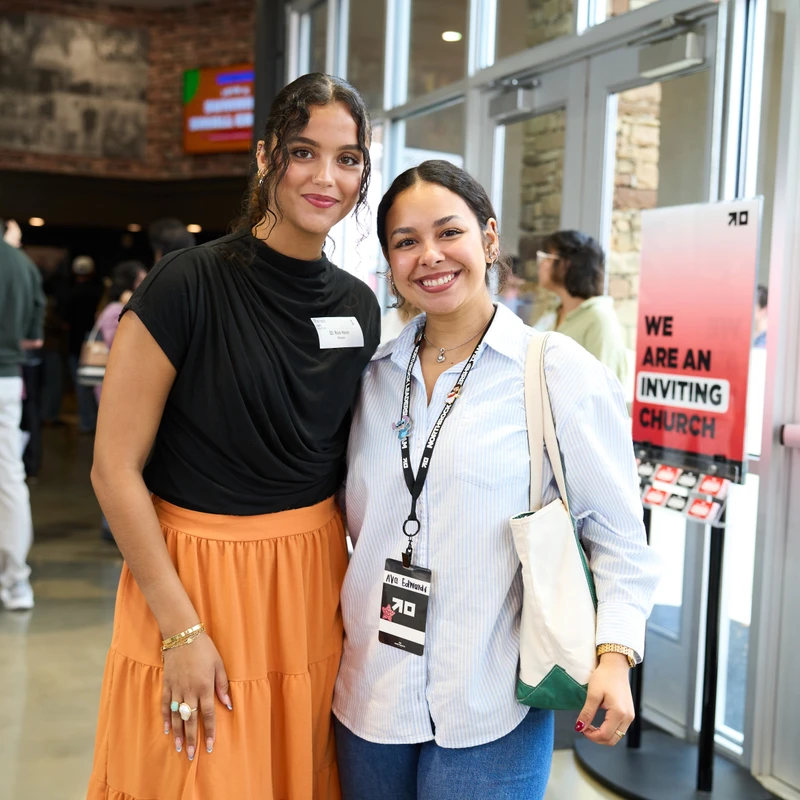 Two smiling women stand side-by-side in a lobby. One wears a black top and orange skirt, the other a striped shirt with a lanyard and a tote bag.